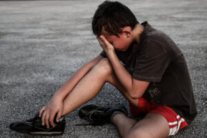 Crying boy sitting down on the pavement with hand covering face